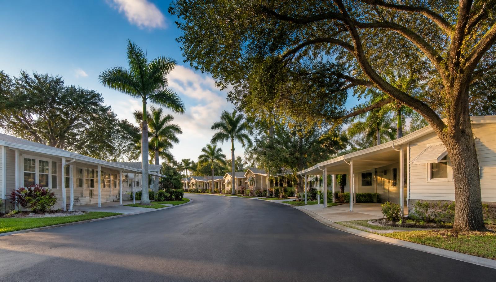 An exterior view of a well maintained South Florida mobile home park and manufactured housing community featuring paved roadways and mature tree canopies.